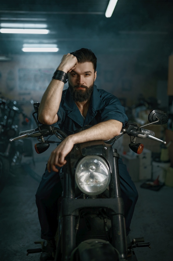 Portrait of brutal unshaven man mechanic sitting on repaired motorcycle over workshop background. Portrait of brutal man mechanic sitting on repaired motorcycle