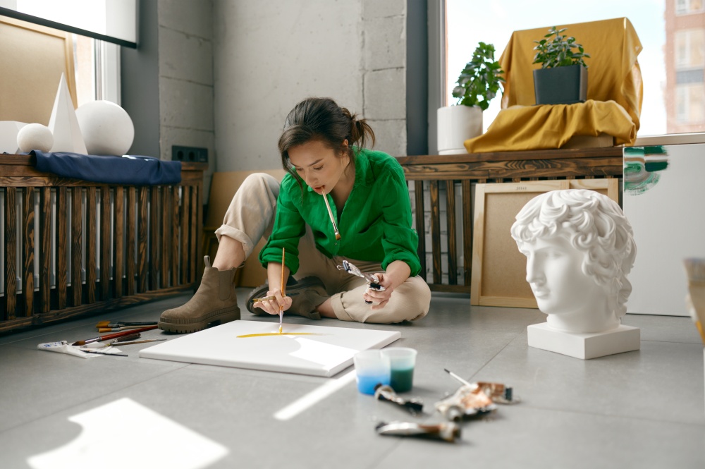 Pretty young female artist drawing picture on canvas with oil paints sitting on floor in her own art studio. Creativity in work and contemporary creative painter concept. Young female artist drawing picture sitting on floor in her own art studio