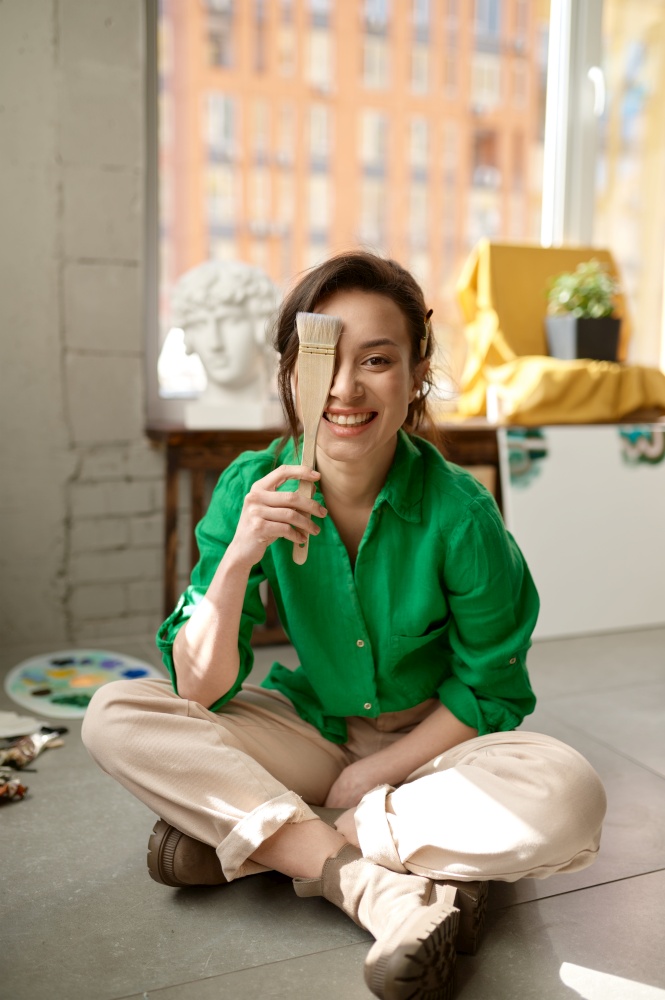 Portrait of creative young woman looking at camera with smile covering her eye with paintbrush. Talented female artist working at home art studio. Portrait of woman looking at camera with smile covering eye with paintbrush