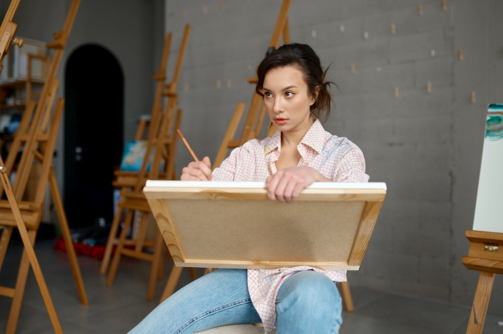 Attractive casual young woman artist holding canvas in hand drawing picture with paints and brush front view. People, leisure and hobby concept. Attractive young woman artist holding canvas in hand drawing picture