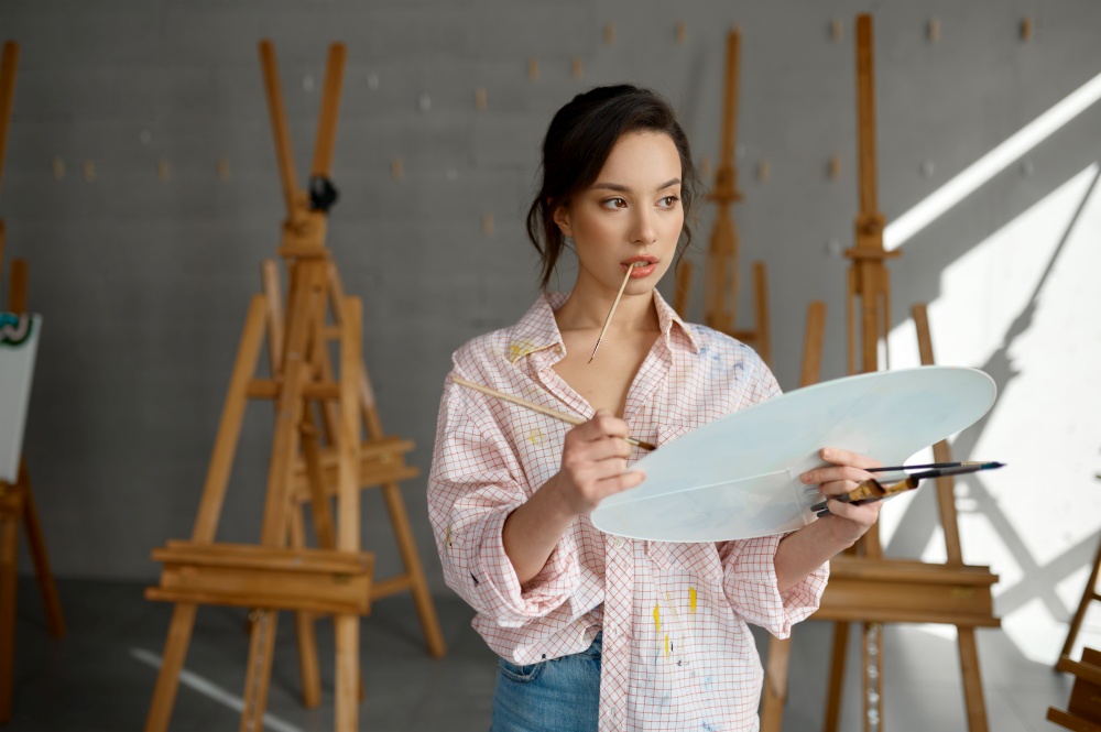 Portrait of thoughtful inspired woman artist holding in hand paints palette looking ahead. Female painter preparing to draw picture mixing colors. Creative art workshop concept. Thoughtful woman artist preparing to draw picture holding in hand paints palette