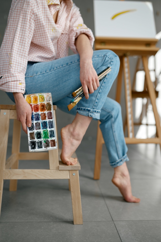 Cropped shot of barefoot female artist holding drawing tools, watercolor paints palette while sitting on chair at home art workspace. Cropped shot of barefoot female artist holding drawing tools sitting on chair