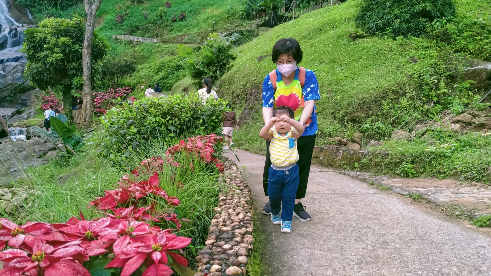 Asian aunt wearing face mask with her little nephew walking on natural walkway amidst greenery and waterfall on the hill, full length view with copy space