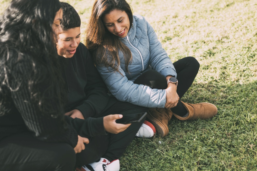 Hispanic family spending quality time together in a local park on a beautiful, sunny day. A teenage boy is sitting on the grass, holding his smartphone while looking away. His mother and sister are sitting next to him, looking at the camera and smiling. Hispanic family spending quality time together in a local park on a beautiful, sunny day. A teenage boy is sitting on the grass, holding his smartphone while looking away. His mother and sister are sitting next to him, looking at the camera and smiling.