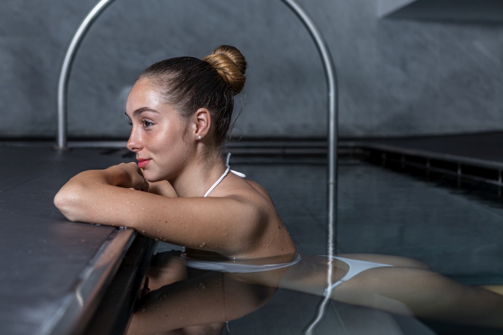 Side view of young female in white bikini with hair bun leaning on pool border and looking away while relaxing in clean water on weekend day on spa resort. Young woman resting in clean pool