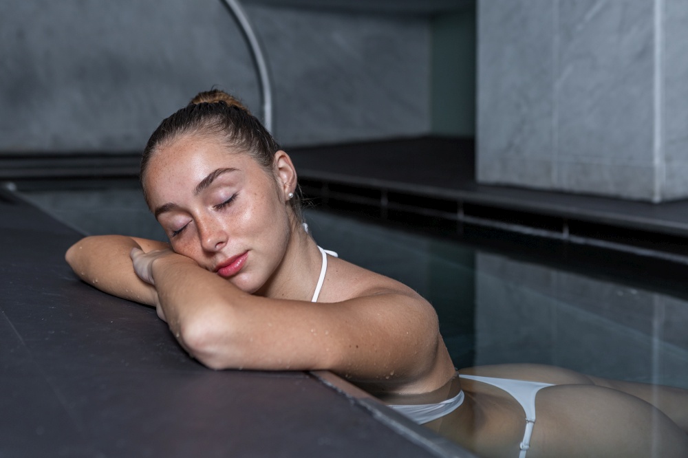 Young female in white bikini closing eyes and holding head on folded arms while chilling in transparent water of clean pool on spa resort. Young woman closing eyes and relaxing in pool