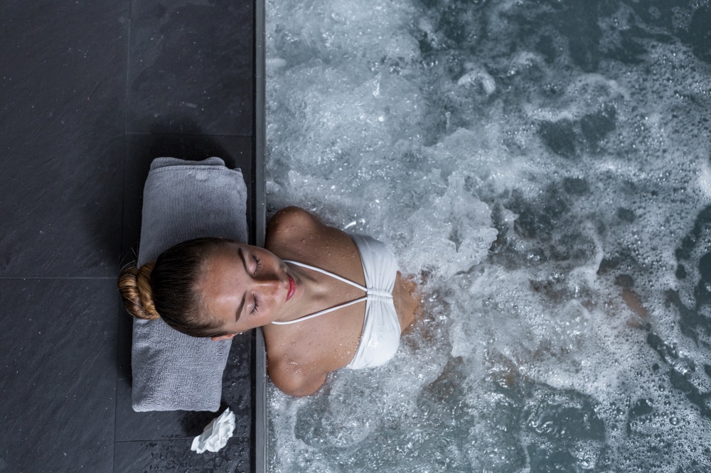 Top view of young woman with closed eyes leaning on pool border with towel and enjoying hydrotherapy in clean pool with splashing water. Young female relaxing in massage pool in spa