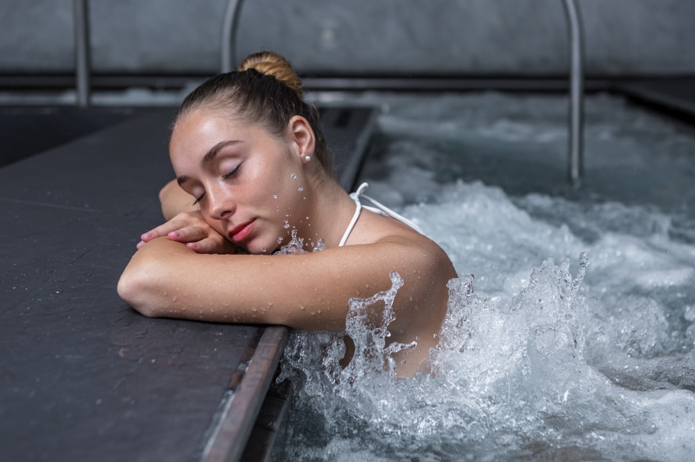 Relaxed young woman closing eyes and holding head on crossed arms while chilling in splashing water of massage pool on weekend day on spa resort. Young female enjoying water massage