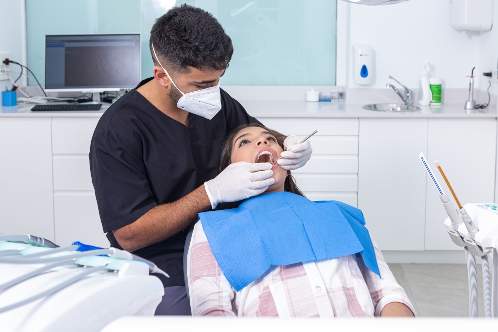 Hispanic male dentist in uniform with gloves and face mask inspecting teeth of teenager during work in modern dental clinic in daytime. Dentist checking teeth of teen patient