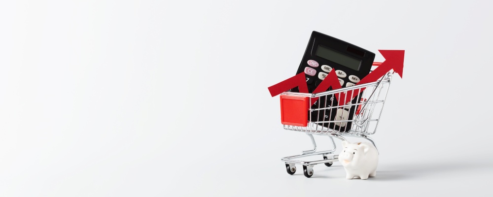 Miniature Shopping cart trolley, calculator, piggy bank and red arrow up on white background with copy space. Concept for grocery expenses and consumerism banner
