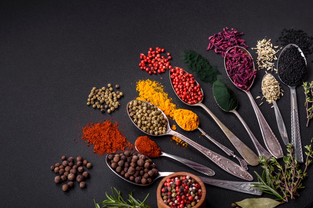 Several types of spices in metal spoons paprika, tomatoes, curry, beets, cumin, turmeric, fennel, spirulina, allspice of different colors on a black concrete background