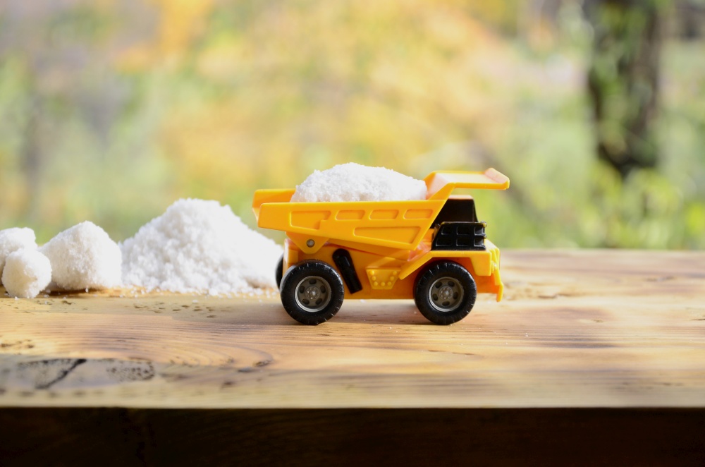A small yellow toy truck is loaded with a stone of white salt next to a pile of salt. A car on a wooden surface against a background of autumn forest. Extraction and transportation of salt