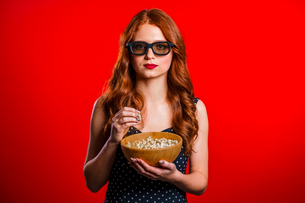 Young woman in 3d glasses watching movie and eating popcorn on red studio background. Young woman in 3d glasses watching movie and eating popcorn on red studio background.