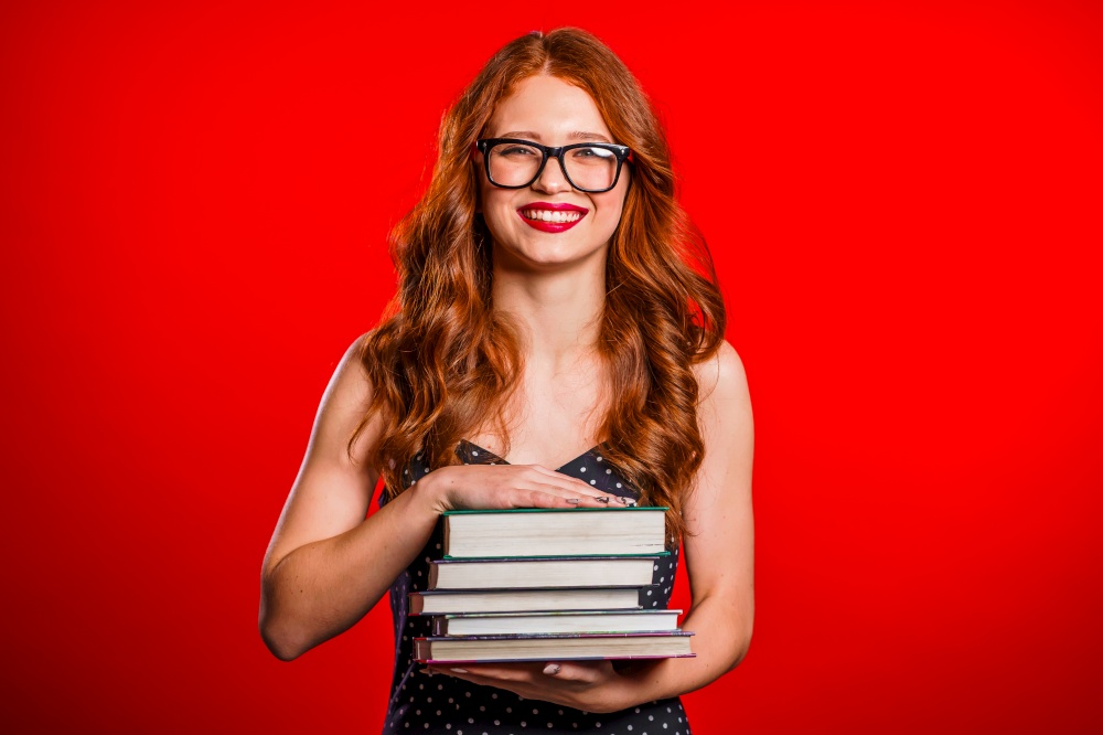 European student on red background in studio holds stack of university books from library. Woman smiles, she is happy to graduate. European student on red background in studio holds stack of university books