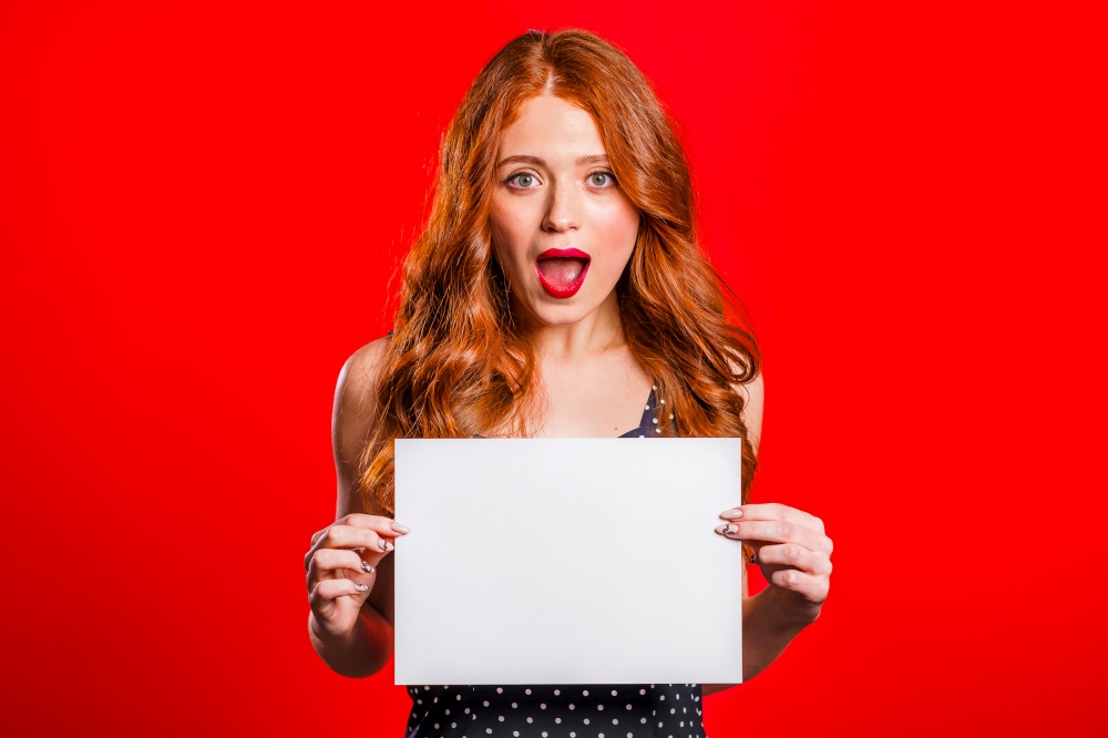 Pretty woman holding white a4 paper poster. Copy space. Smiling trendy girl with red hair on studio background. Pretty woman holding white a4 paper poster.Copy space.Smiling girl with red hair
