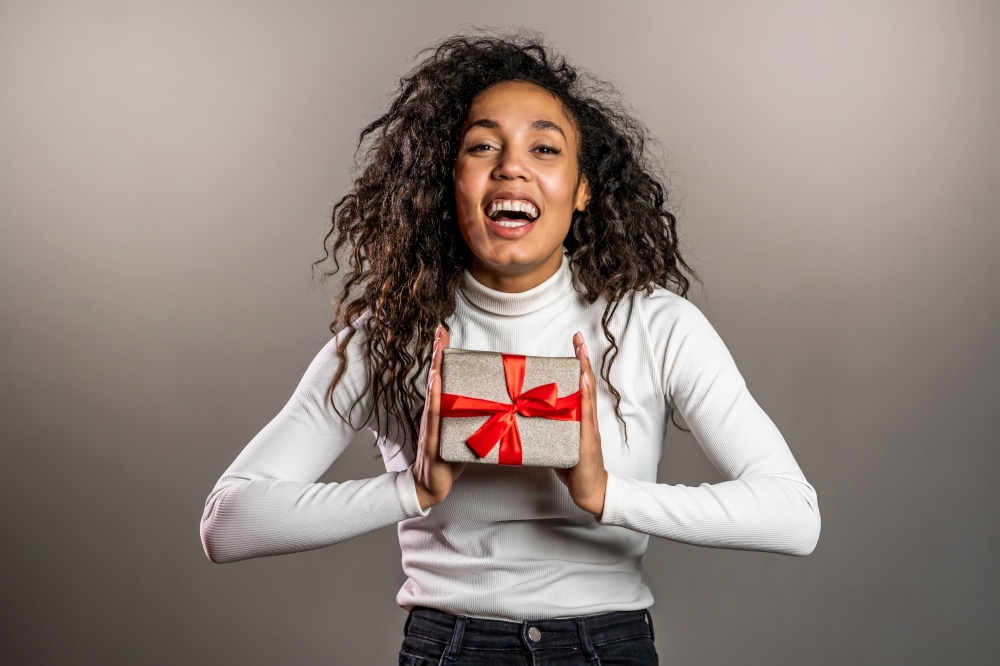 Pretty birthday woman curly hair with gift box on grey background. African american lady smiling, she is happy with present. High quality photo. Pretty birthday woman curly hair with gift box on grey background. African american lady smiling, she is happy with present.