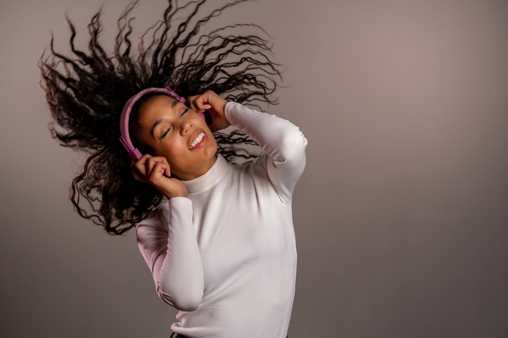 Pretty mixed race girl with curly afro hair having fun, smiling, dancing with headphones in studio on grey background. Music, dance, radio concept. Pretty mixed race girl with curly afro hair having fun, smiling, dancing with headphones in studio on grey background. Music, dance, radio concept.