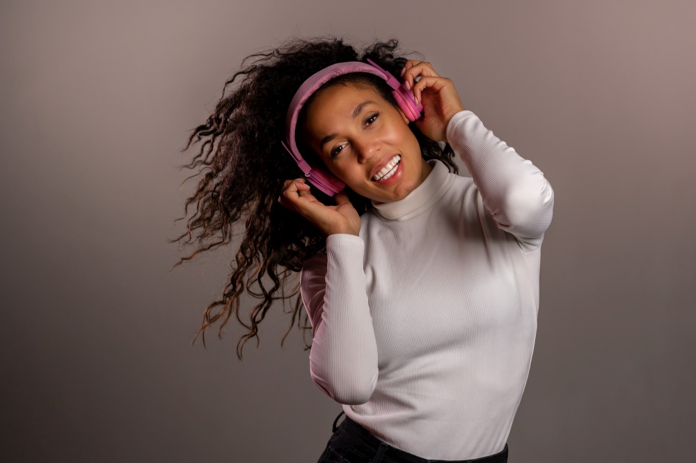 Pretty mixed race girl with curly afro hair having fun, smiling, dancing with headphones in studio on grey background. Music, dance, radio concept. Pretty mixed race girl with curly afro hair having fun, smiling, dancing with headphones in studio on grey background. Music, dance, radio concept.