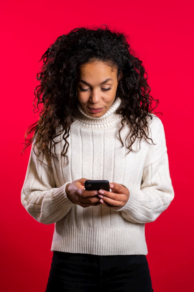 Girl with afro hair using smartphone, surfing internet or playing game on red studio background. Modern technology - apps, social networks.. Girl with afro hair using smartphone, surfing internet or playing game on red studio background. Modern technology - apps, social networks