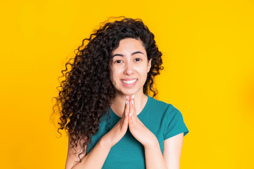 Attractive spanish woman in green t-shirt praying over yellow background. Mixed race girl begging someone. Attractive spanish woman in green t-shirt praying over yellow background. Mixed race girl begging someone.