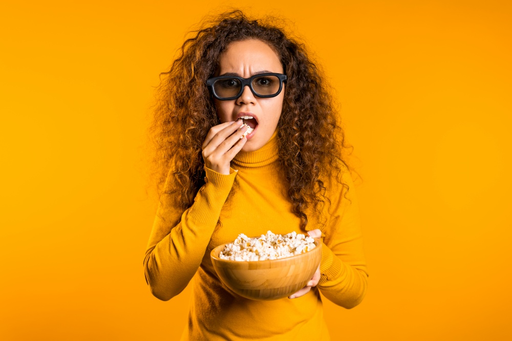 Young woman in 3d glasses watching movie and eating popcorn on yellow studio background. Young woman in 3d glasses watching movie and eating popcorn on yellow background