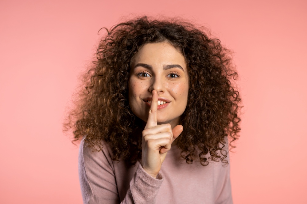 Positive girl holding finger on her lips over pink background. Gesture of shhh, secret, silence. Close up. Body language. Young curly woman with trendy glitter freckles make-up.. Positive smiling girl holding finger on her lips over pink background. Gesture of shhh, secret, silence. Close up. Body language. Young curly woman with trendy glitter freckles make-up