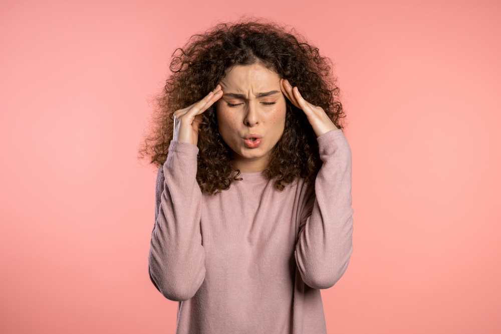 Young beautiful woman with curly hair having headache, pink studio portrait. Girl putting hands on head, isolated on background. Concept of problems and headache. Young beautiful woman with curly hair having headache, pink studio portrait. Girl putting hands on head, isolated on background. Concept of problems and headache.