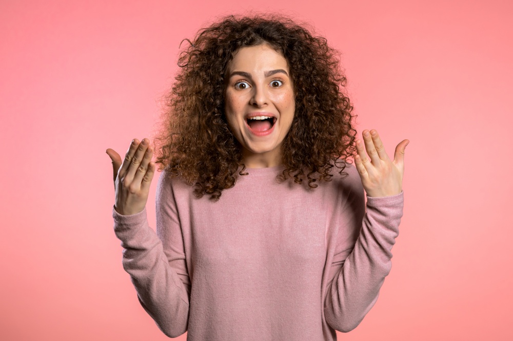 Portrait of girl with curly hair, she shows wow delight effect gesture. Surprised excited happy woman. Pretty female shocked model on pink background. Portrait of girl with curly hair, she shows wow delight effect gesture. Surprised excited happy woman. Pretty female shocked model on pink background.