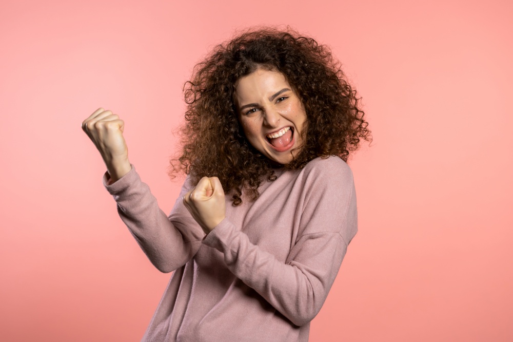 Girl with curly hair and extraordinary makeup shows yes gesture of victory, she achieved result, goals. Surprised excited happy woman on pink background.. Girl with curly hair and extraordinary makeup shows yes gesture of victory, she achieved result, goals. Surprised excited happy woman on pink background