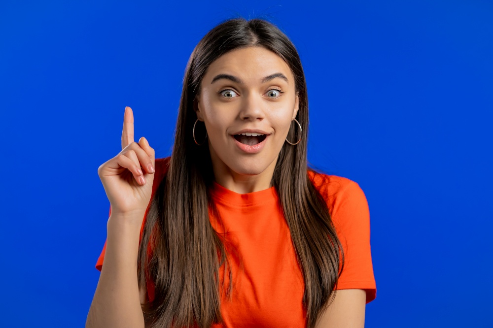 Portrait of young thinking pondering woman having idea moment pointing finger up on blue studio background. Smiling happy girl showing eureka gesture. Portrait of young thinking pondering woman having idea moment pointing finger up on blue studio background. Smiling happy girl showing eureka gesture.