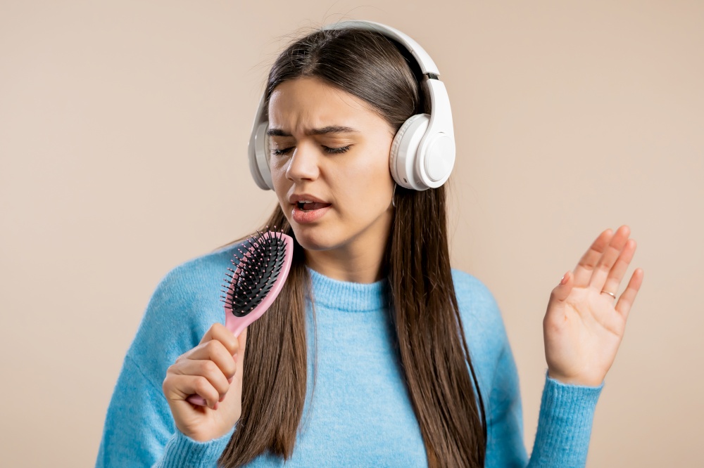 European woman singing and dancing with hair brush or comb instead microphone at studio background. Lady in headphones having fun, listening to music, dreams of being celebrity. High quality. European woman singing and dancing with hair brush or comb instead microphone at light studio background. Lady in headphones having fun, listening to music, dreams of being celebrity.