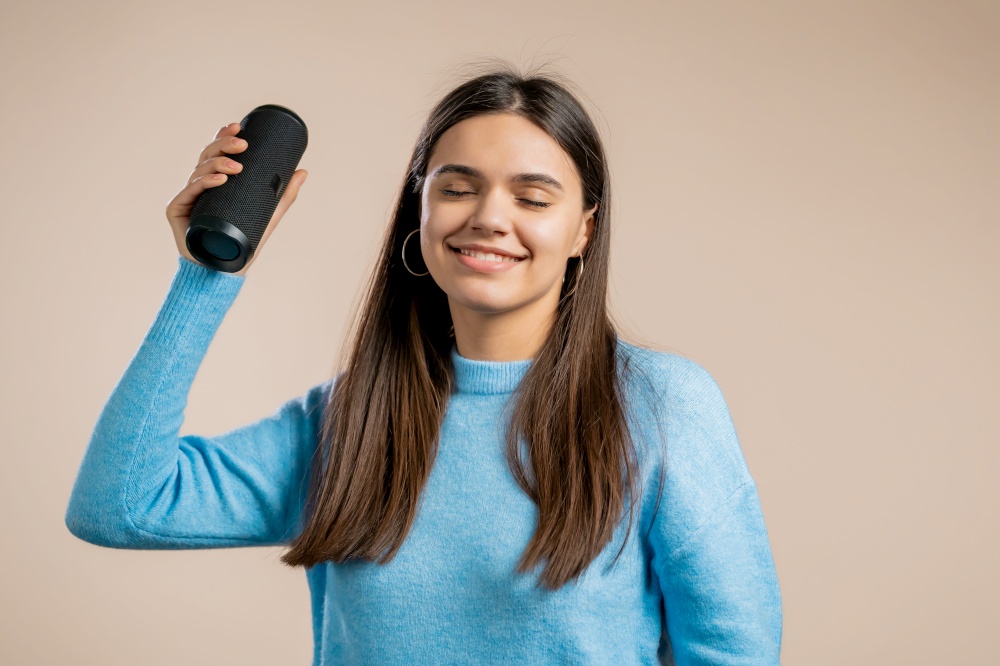 Young woman listening to music by wireless portable speaker - modern sound system. Lady dancing, enjoying on beige studio background. She moves to the rhythm of music. High quality. Young woman listening to music by wireless portable speaker - modern sound system. Lady dancing, enjoying on beige studio background. She moves to the rhythm of music.