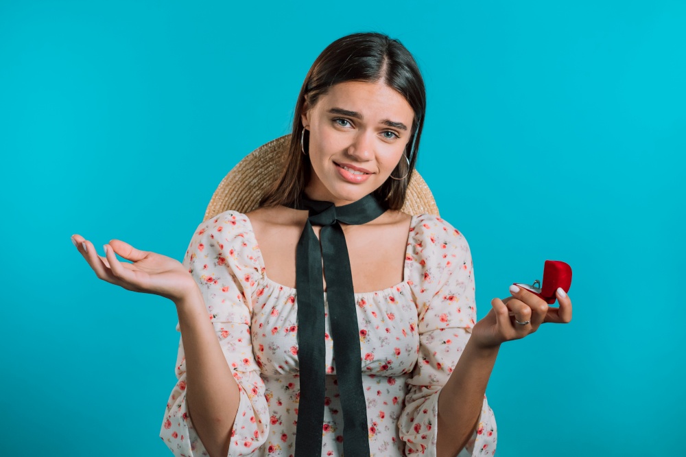 Pretty european woman in floral dress holding small jewelry box with marriage proposal on blue background. Disappointed upset girl, she doesn&rsquo;t like gift. . Disappointed upset girl, she doesn&rsquo;t like gift. Pretty european woman in floral dress holding small jewelry box with marriage proposal on blue background.