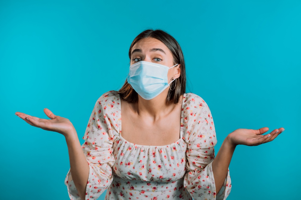 Unsure woman in medical mask shrugs her arms, makes gesture of I don&rsquo;t know, care, can&rsquo;t help anything . Girl on blue background.. Unsure woman in medical mask shrugs her arms, makes gesture of I don&rsquo;t know, care, can&rsquo;t help anything . Girl on blue background
