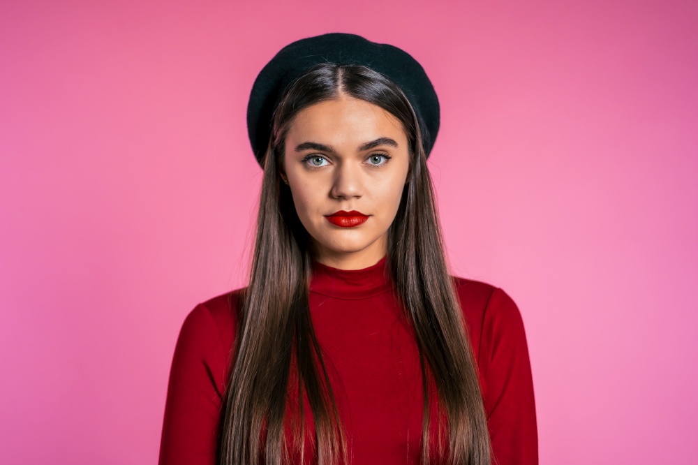 Beautiful cheerful european woman in red wear and french hat smiling to camera over pink wall background. Cute trendy girl&rsquo;s portrait. Beautiful cheerful european woman in red wear and french hat smiling to camera