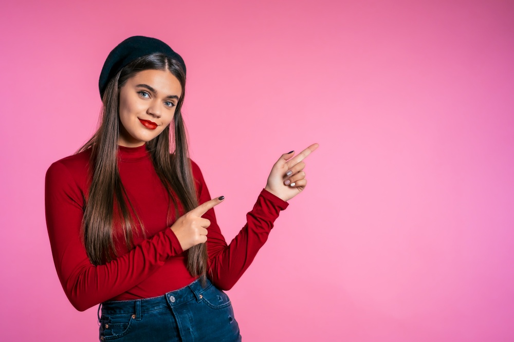 Happy smiling woman in red wear presenting and showing something isolated on pink background. Portrait of girl, she pointing with arms on her left with copy space. Happy smiling woman in red wear presenting showing something isolated on pink