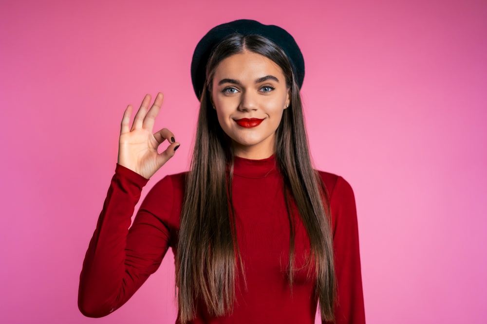 Winner. Success. Positive girl making OK sign over pink background and smiles to camera. Body language. Young hipster woman with perfect red lips make-up and french hat. Winner. Success. Positive girl making OK sign over pink background and smiles