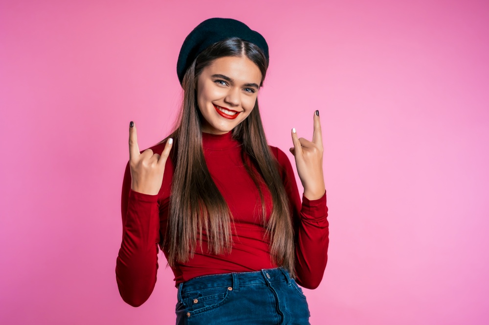 Girl with long hair and pretty appearance shows rock sign. Smiling excited happy hipster on pink background.. Girl with long hair and pretty appearance shows rock sign.