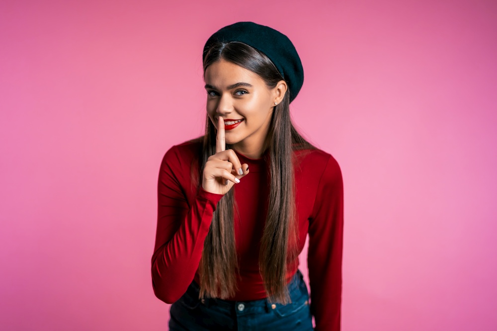 Beautiful woman with red lips holding finger on her lips over pink background. Gesture of shhh, secret, silence. Close up. Beautiful woman with red lips holding finger on her lips over pink background.
