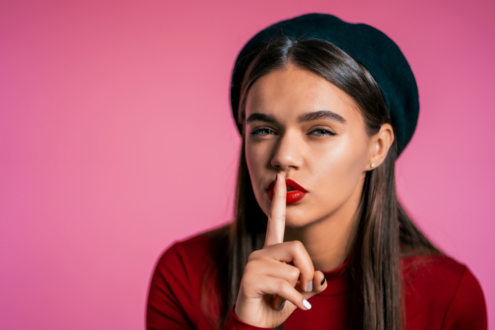 Beautiful woman with red lips holding finger on her lips over pink background. Gesture of shhh, secret, silence. Close up. Beautiful woman with red lips holding finger on her lips over pink background.
