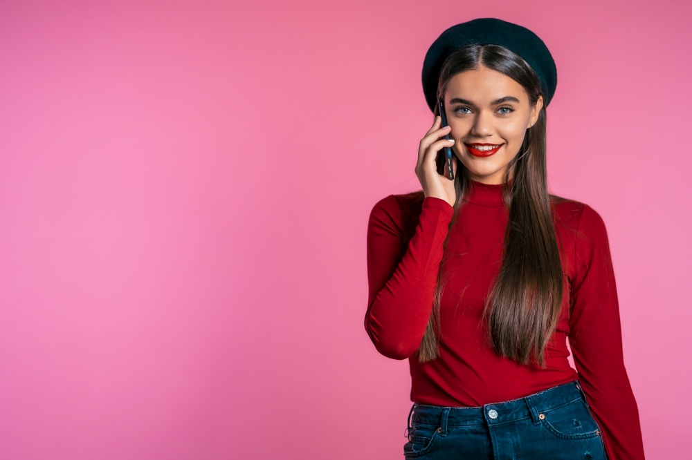 Smiling hipster talking on mobile phone over pink background. Beautiful woman holding and using smart phone. Girl with technology concept. Copy space. Smiling hipster talking on mobile phone over pink background. Beautiful woman