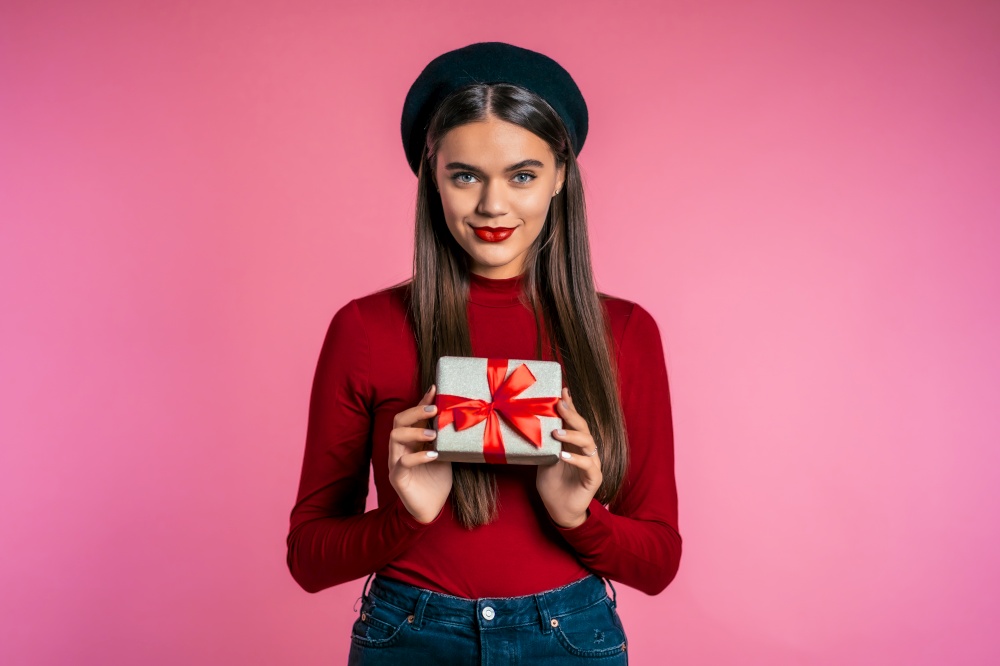Joyful beautiful woman with perfect makeup holding gift box with bow on pink wall background. Trendy girl smiling, she is glad to get present. Joyful beautiful woman with perfect makeup holding gift box with bow on pink