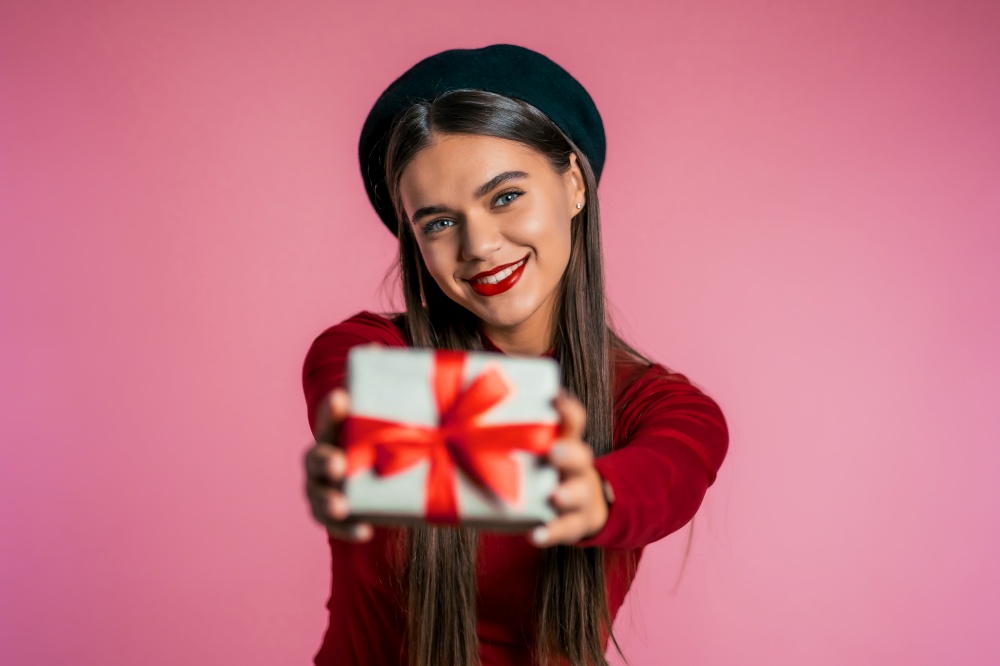 Pretty woman gives gift and hands it to the camera. She is happy, smiling. Girl on pink background. Positive holiday shot. Pretty woman gives gift and hands it to the camera. She is happy, smiling.