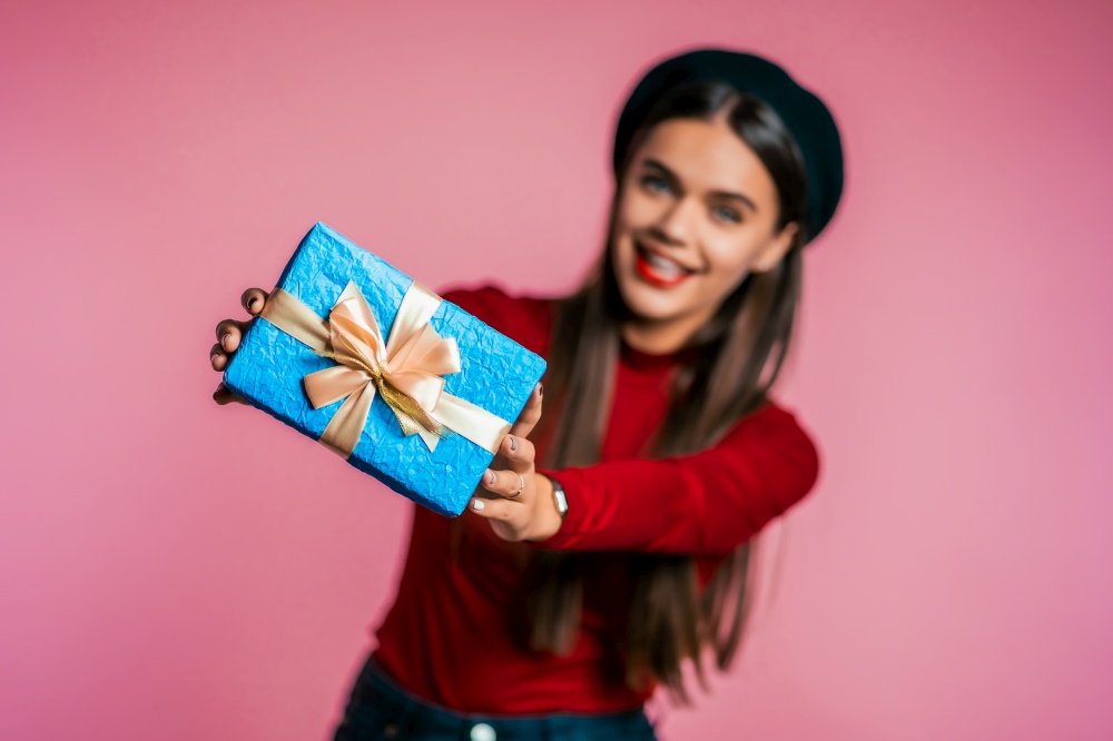 Pretty woman gives gift and hands it to the camera. She is happy, smiling. Girl on pink background. Positive holiday shot. Pretty woman gives gift and hands it to the camera. She is happy, smiling. Girl on pink background. Positive holiday shot.