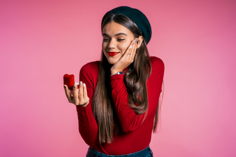 Pretty european young woman in red holding small jewelry box with proposal diamond ring on pink wall background. Girl smiling, she is happy to get present, proposition for marriage. Pretty european young woman in red holding small jewelry box with proposal ring