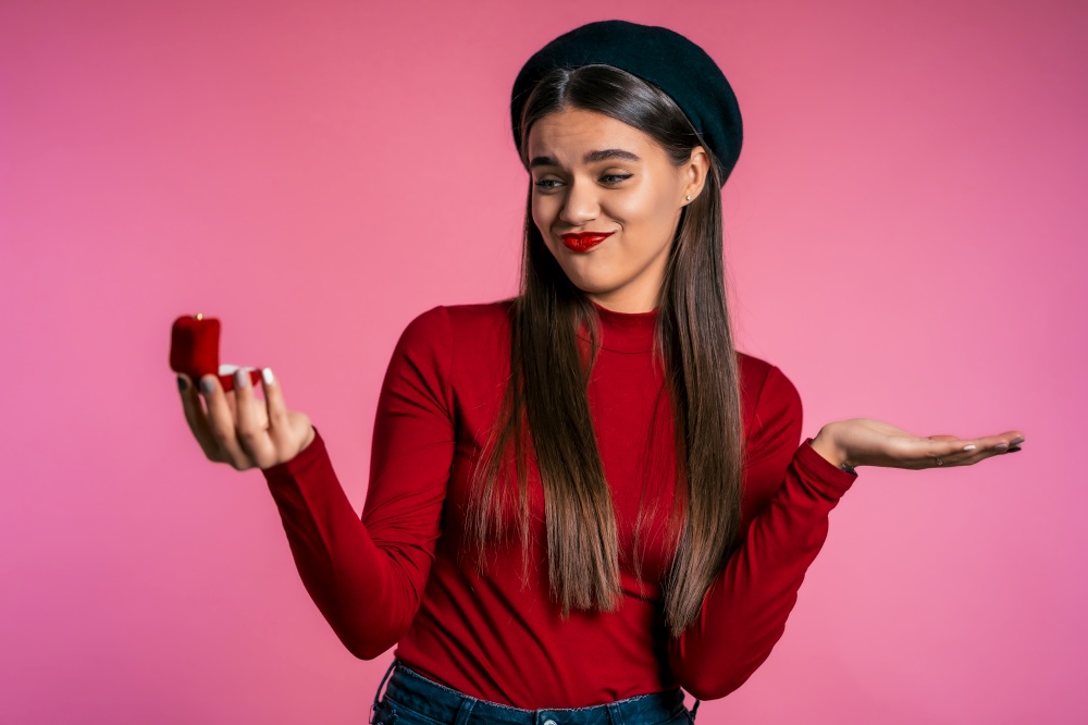 Pretty european young woman in red holding small jewelry box with proposal diamond ring on pink wall background. Disappointed upset girl, she doesn&rsquo;t like gift. Disappointed upset girl, she doesn&rsquo;t like gift