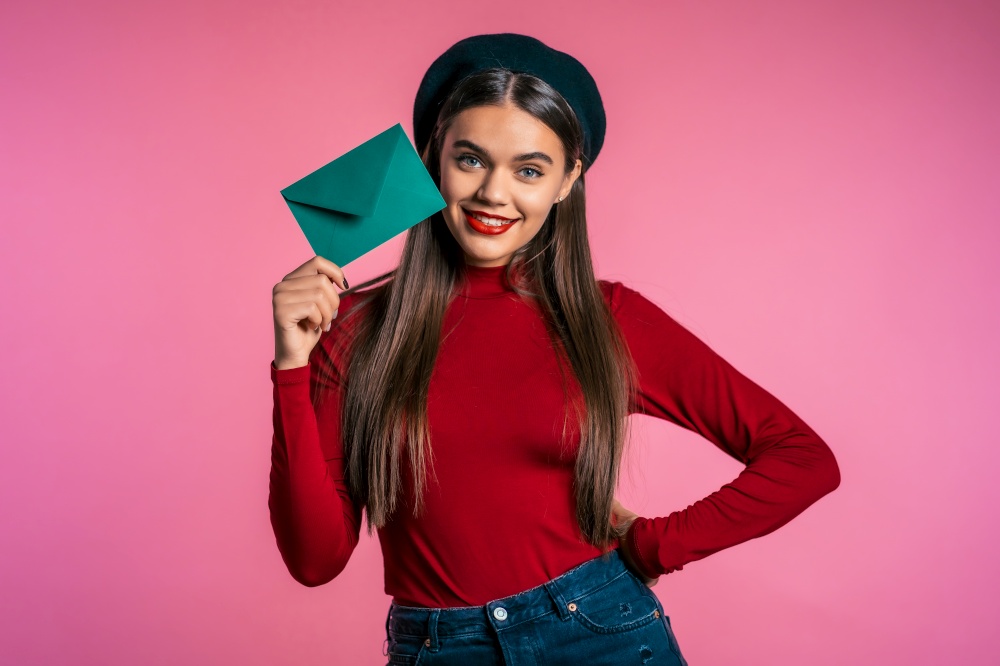 Portrait of young beautiful woman in red wear and hat with green envelope on pink studio background. Portrait of young beautiful woman red wear and hat with green envelope on pink