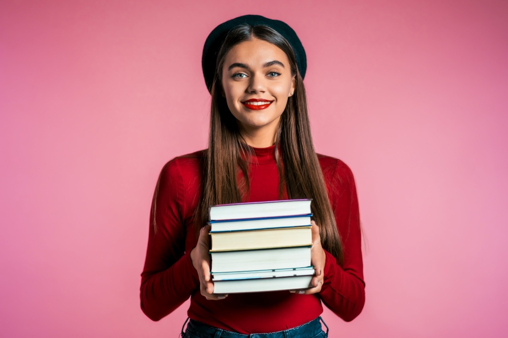 Pretty student on pink background in studio holds stack of university books from library. Girl in hat smiles, she is happy to graduate. student pink background in studio holds stack of university books from library
