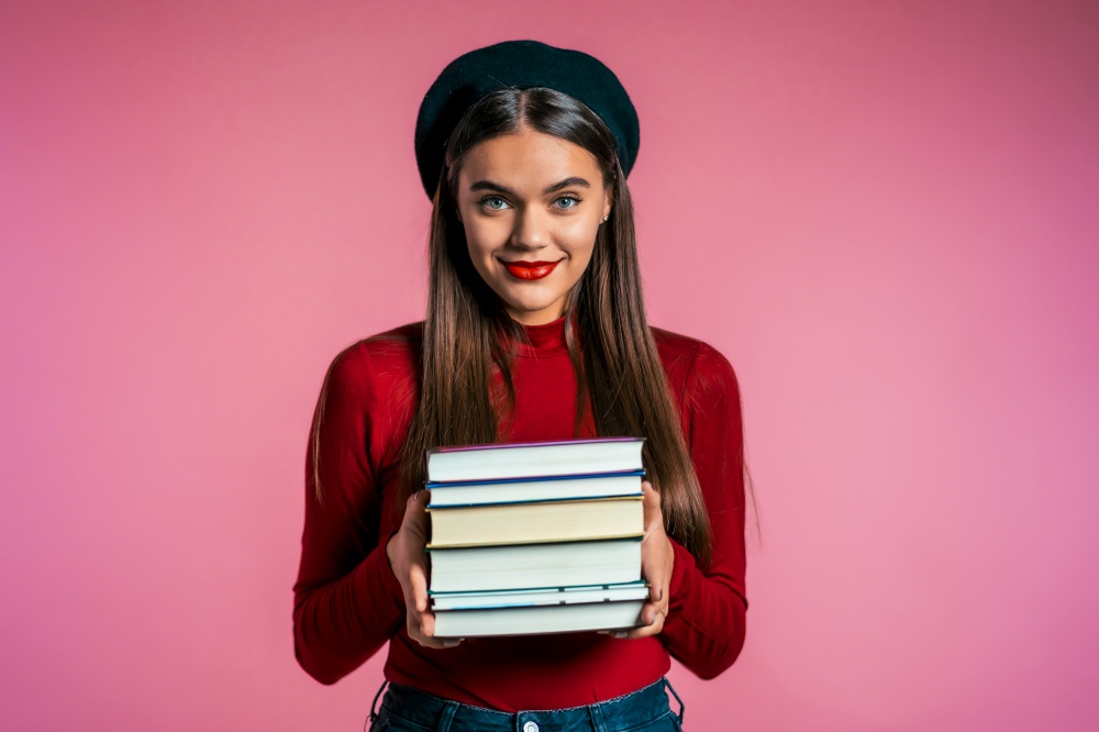 Pretty student on pink background in studio holds stack of university books from library. Girl in hat smiles, she is happy to graduate. student pink background in studio holds stack of university books from library