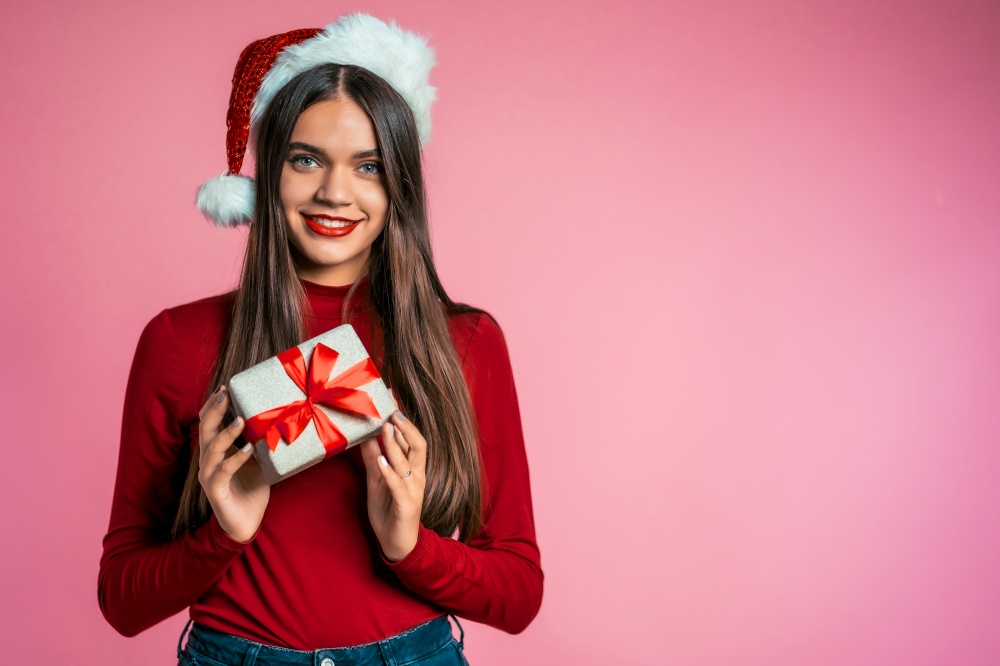 Attractive girl in Santa hat with gift box. Portrait of lady on pink background. Christmas or New year concept. Happy pretty woman smiling to camera. Attractive girl in Santa hat with gift box. Portrait of lady on pink background.